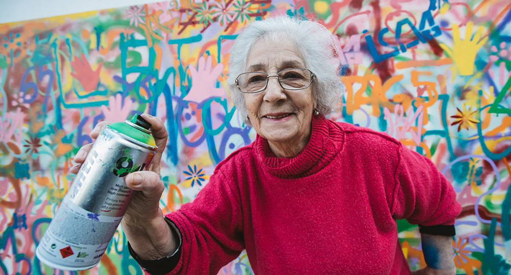 An older woman with white hair, wearing a red sweater, holding a spray paint can.