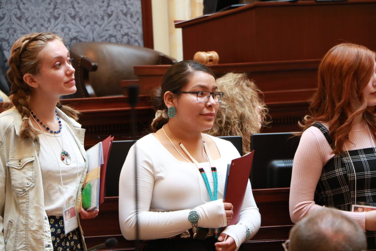 Three young people standing on the Utah Senate floor holding folders.