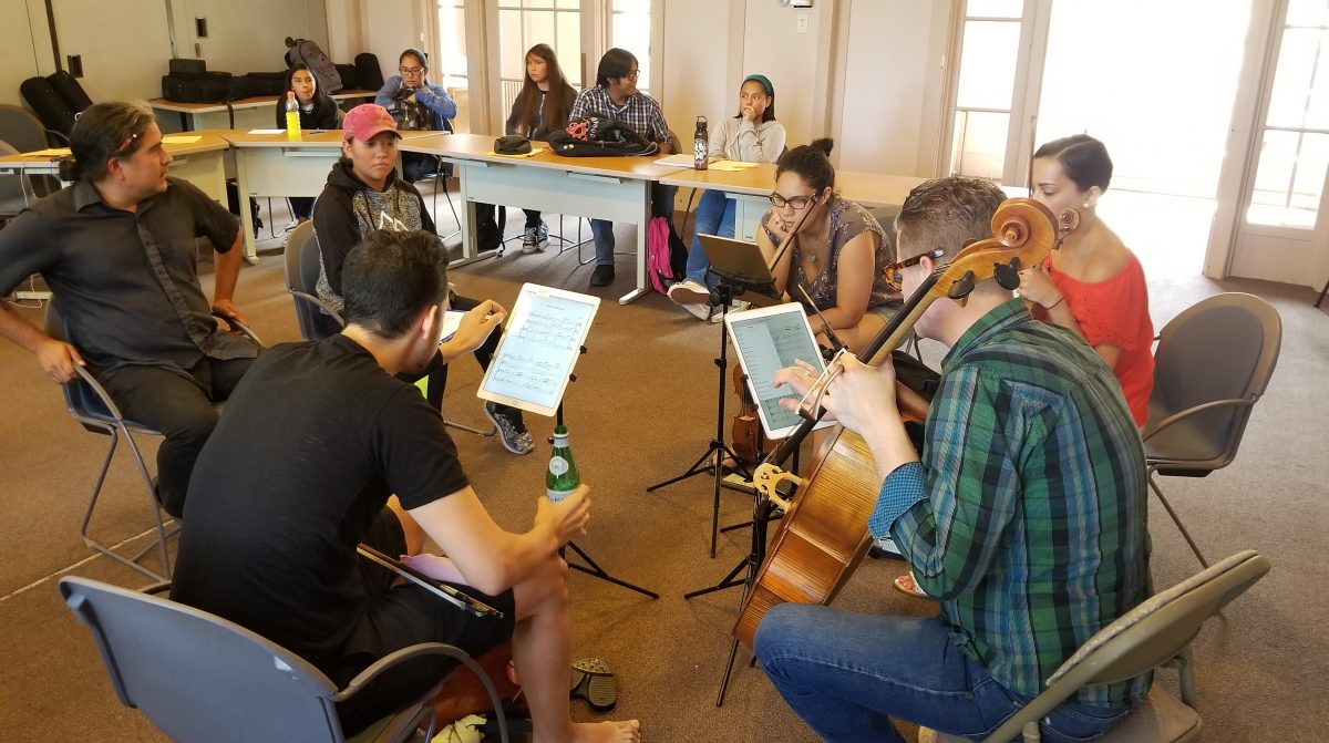 An image of musicians sitting in a circle reading sheet music.