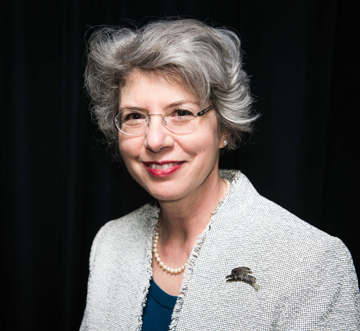 Portrait of a woman with grey hair smiling, wearing glasses and a grey blazer.