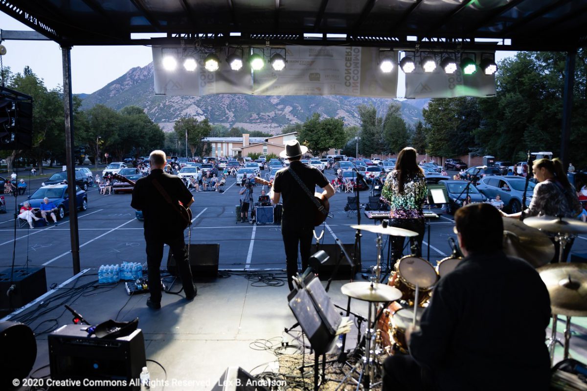 A band plays on a stage at a parking lot concert. Viewers are spread apart sitting in or around their cars.