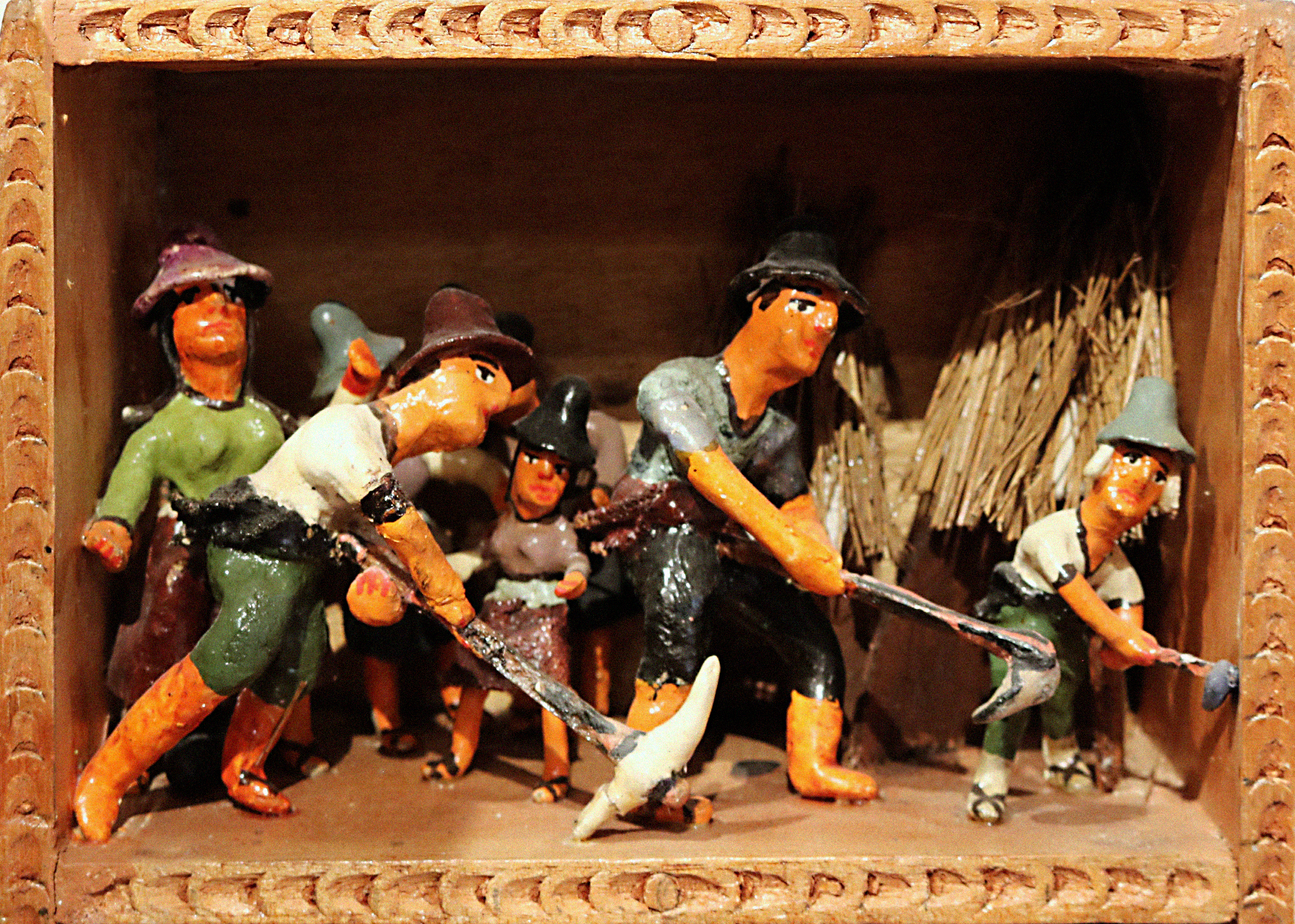 A scene called Sowing within a retablo. The scene portrays humanlike figurines wearing hats and holding farming tools, as though they are working in a field. In the backgroup are what appear to be structures with straw roofs.