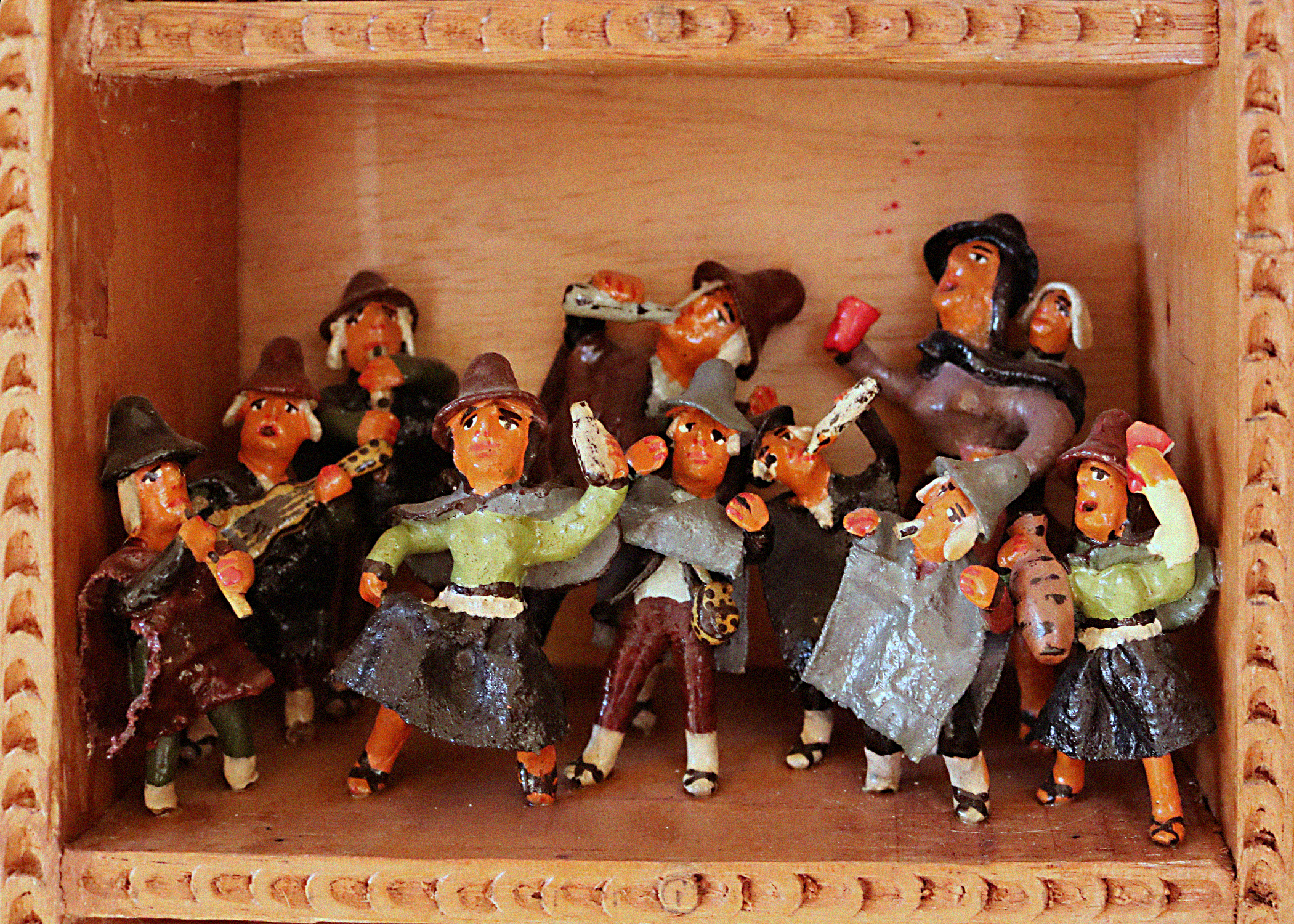 A scene called Traditional Customs within a retablo. The scene portrays humanlike figurines in a group. The men and women portrayed wear earthy colors like green, grey, brown, and dark red. All the figurines wear hats. Most hold and drink from white bottles. One figurine holds a guitar and two other hold flutes to their mouths.