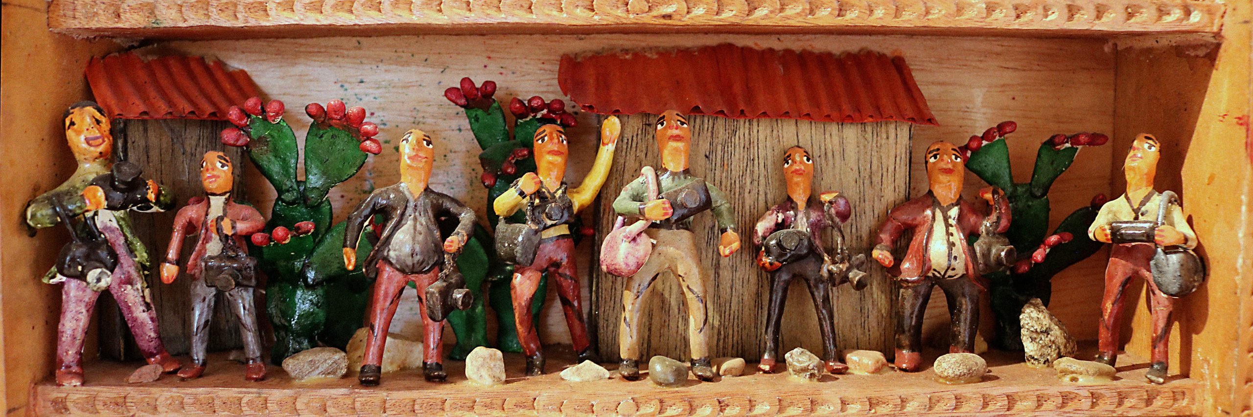 A scene called The Martyrs of Journalism within a retablo. The scene portrays eight men standing together holding bags and cameras. The men stand in from of the sides of two buildings with wooden walls and red, tile roofs. In the background are also three green cactuses with red flowers. The group around the men is scattered with rocks.