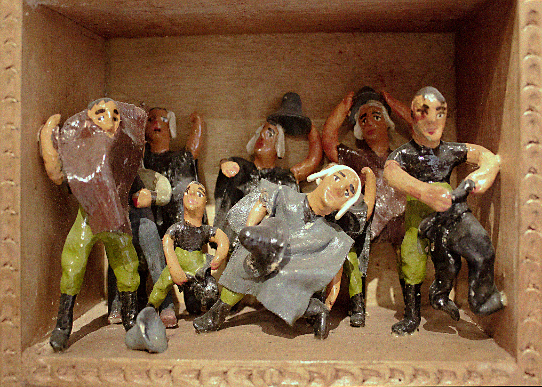 A scene called Disguise within a retablo. A group of nine humanlike figurines are portrayed as dressing themselves in grey and brown ponchos and grey hats.