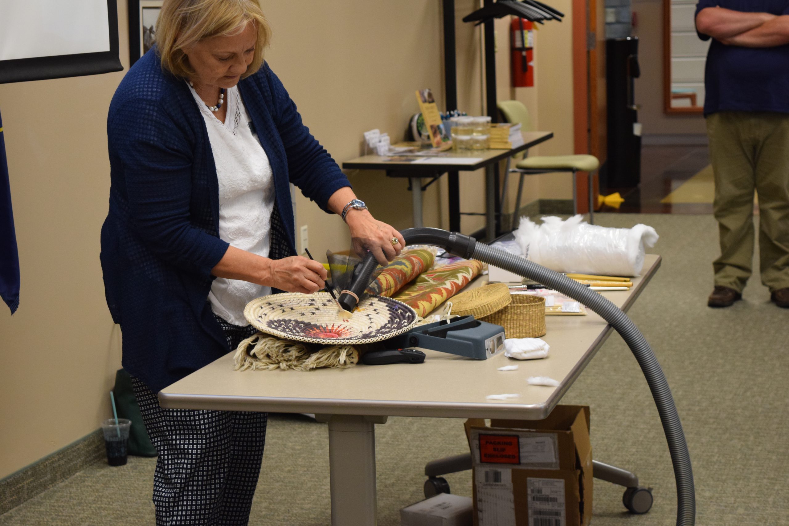 A blonde woman in a blue cardigan cleans a Navajo basket in a collections care workshop.