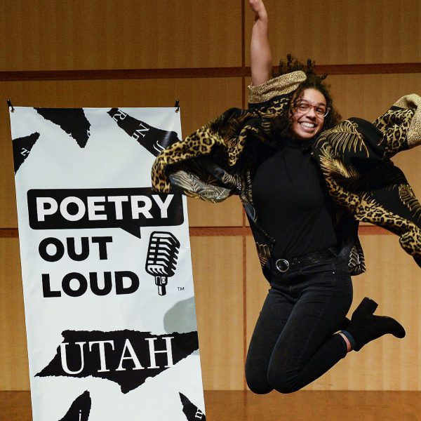 A girl wearing all black with curly black hair jumps and poses next to a sign that says, "Poetry Out Loud Utah."