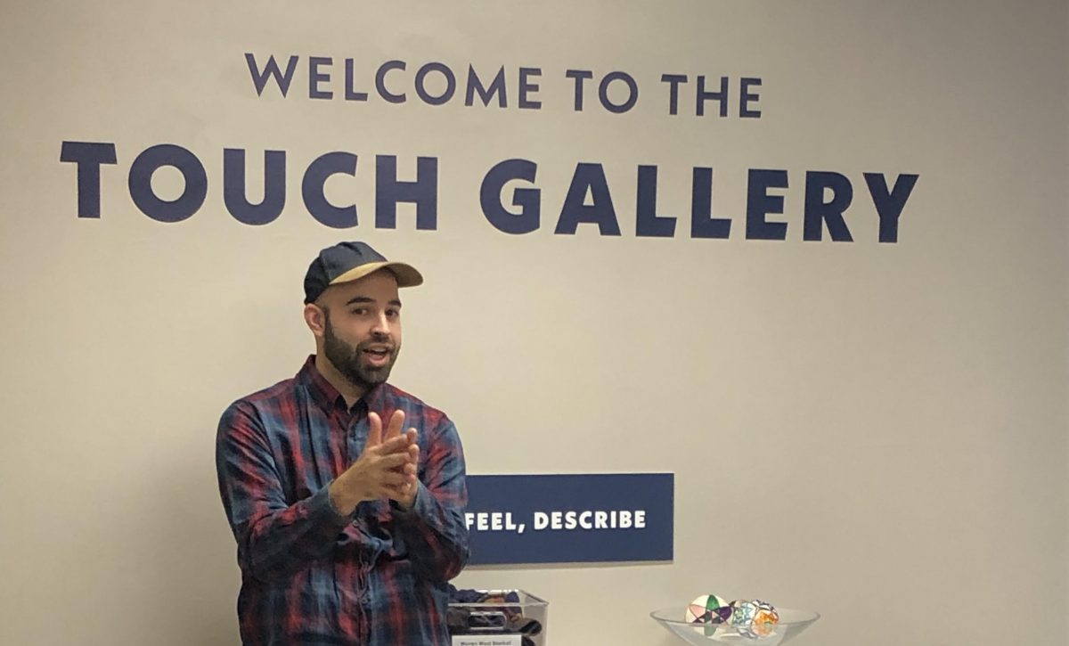 A bearded man in a plaid shirt and ball cap speaks in front of a sign that reads, "Welcome to the Touch Gallery."