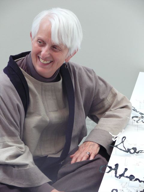 A woman with short, gray hair sits next to an artwork.