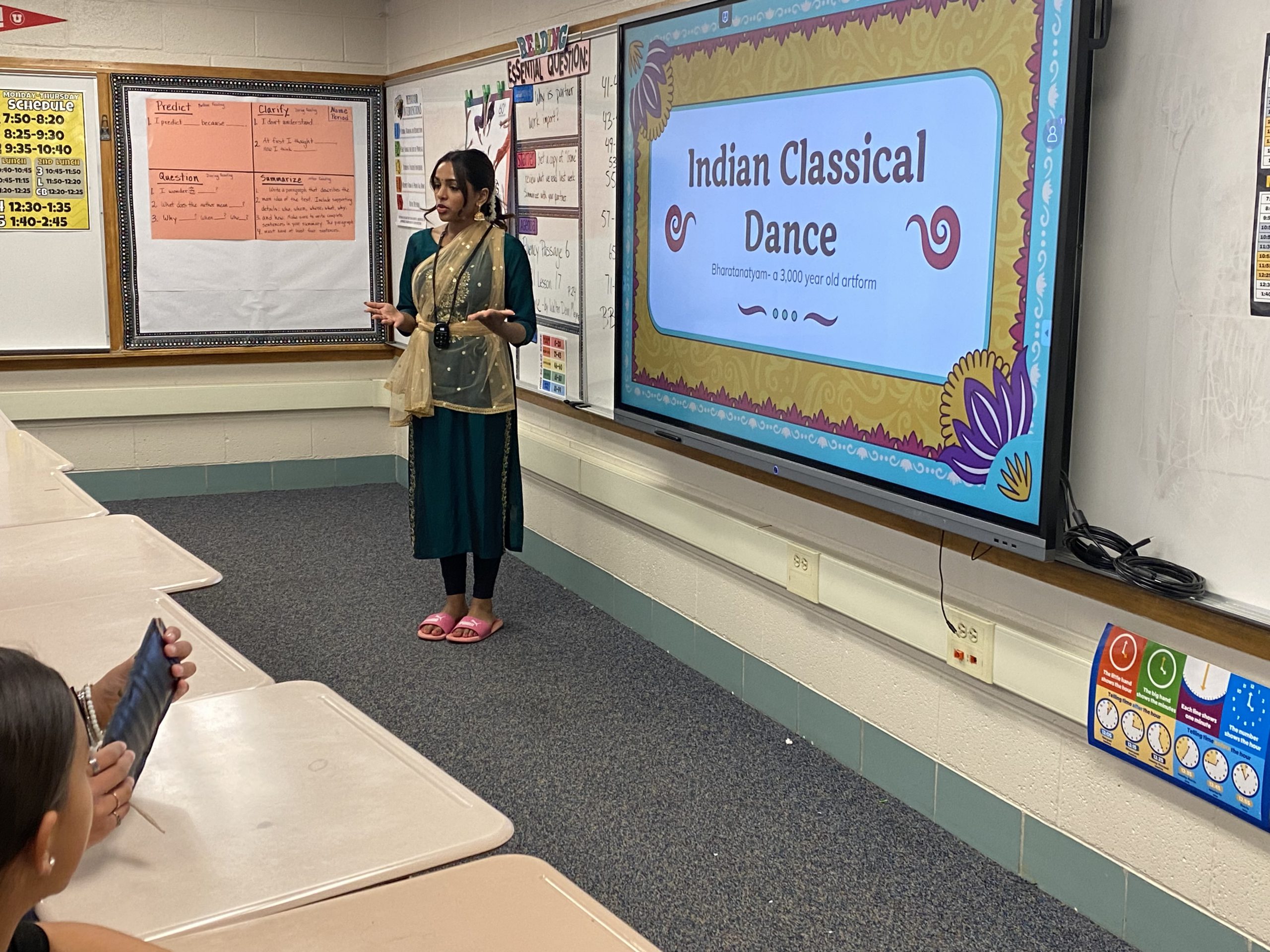 Teacher stands in front of a classroom with a slideshow about Indian dance