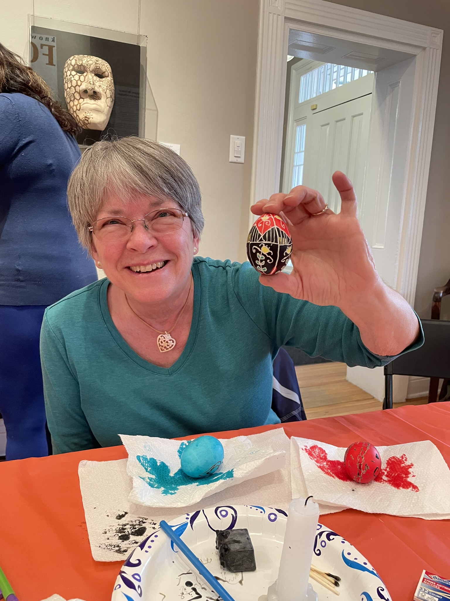 Smiling woman holds up her decorated egg, supplies on table beneath her